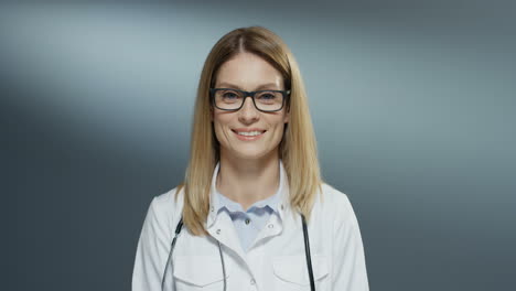 Portrait-shot-of-the-young-Caucasian-beautiful-blonde-woman-doctor-in-glasses-and-white-medical-gown-smiling-happily-to-the-camera.-Close-up.